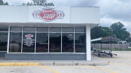 Exterior of a BBQ restaurant with large windows, featuring a sign that reads "BBQ" and a trailer parked nearby.