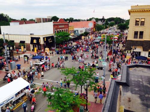 A bustling street filled with crowds, shops, and trees, capturing a lively community event in an urban setting.