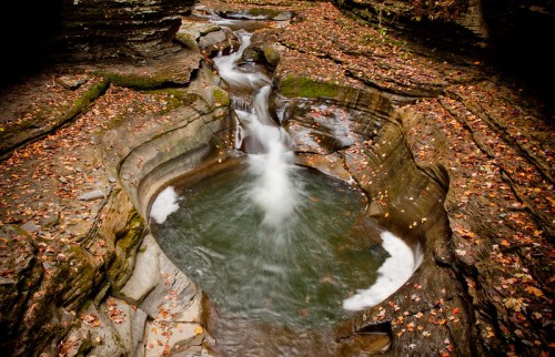 A serene stream flows through rocky terrain, surrounded by autumn leaves and smooth stone formations.