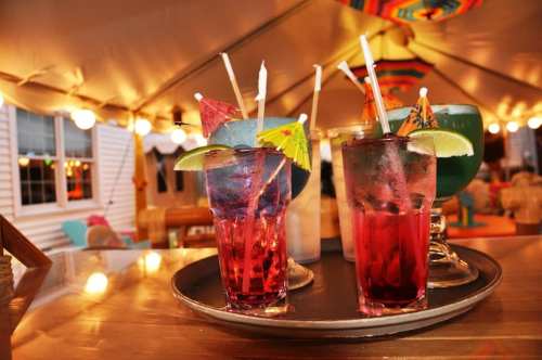 A tray of colorful cocktails with umbrellas and garnishes, set under a festive tent with warm lighting.