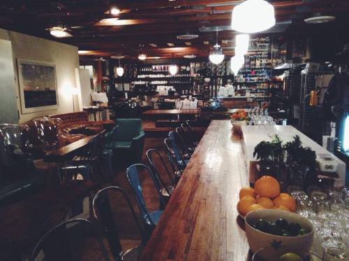 A cozy bar interior with wooden counters, colorful chairs, and shelves stocked with bottles in a warm, inviting atmosphere.