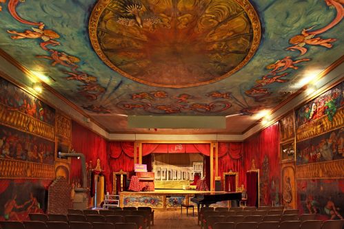 A vibrant theater interior with ornate murals, a painted ceiling, red curtains, and a grand piano on stage.