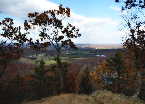 A scenic view from a mountain, featuring trees and a landscape of rolling hills and valleys under a cloudy sky.