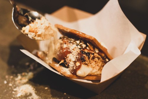 A close-up of a dessert sandwich being topped with crushed nuts, served in a paper tray.