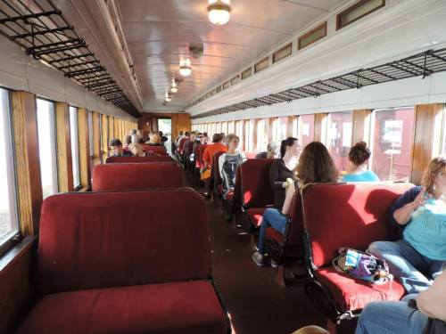 Interior of a vintage train car with rows of red seats and passengers seated along the aisle, enjoying the ride.