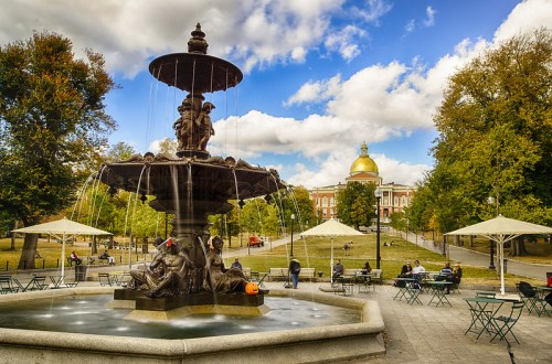 A decorative fountain in a park with a golden dome building in the background and people sitting at tables.