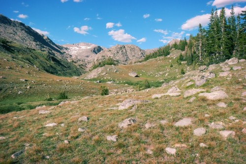 A scenic mountain landscape with rocky terrain, green grass, and trees under a blue sky with scattered clouds.