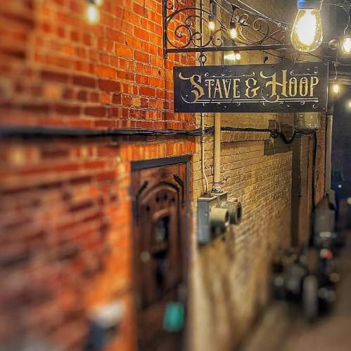 A dimly lit alleyway featuring a vintage sign that reads "Stave & Hoop," with exposed brick walls and hanging lights.