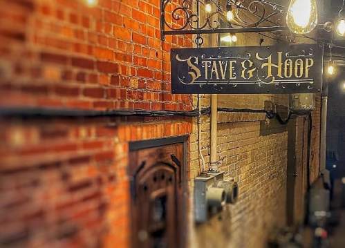 Sign for "Stave & Hoop" hanging in a brick alleyway, illuminated by warm string lights.