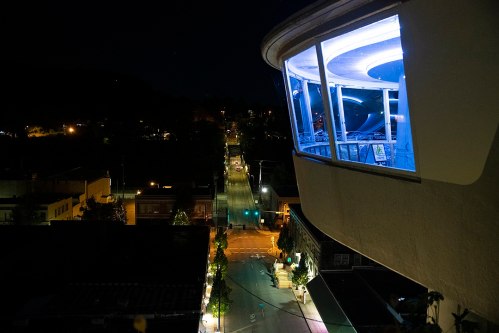 A nighttime view of a street lined with trees, featuring a modern building with illuminated windows.