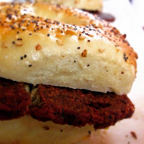 Close-up of a bagel sandwich with a crispy, seasoned patty and a poppy seed topping.