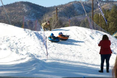 A snowy hill with two people on inflatable tubes, while another person takes a photo nearby. Mountains in the background.