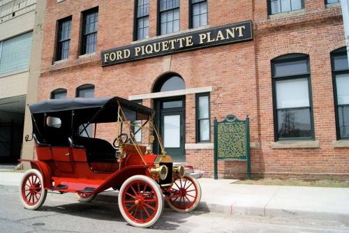 A vintage red car parked in front of the historic Ford Piquette Plant building with a brick facade.