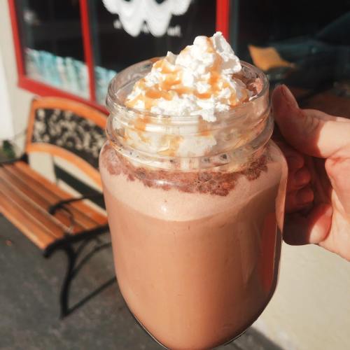 A hand holding a mason jar filled with chocolate drink topped with whipped cream and caramel drizzle, with a bench in the background.