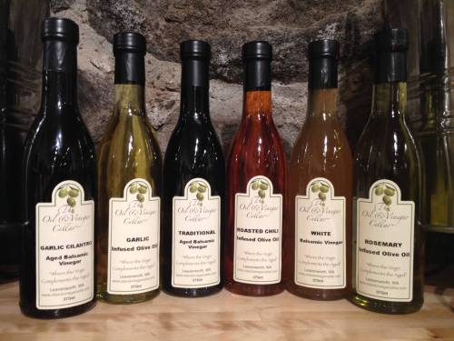 A variety of six bottles of infused olive oils and aged balsamic vinegars on a wooden surface.