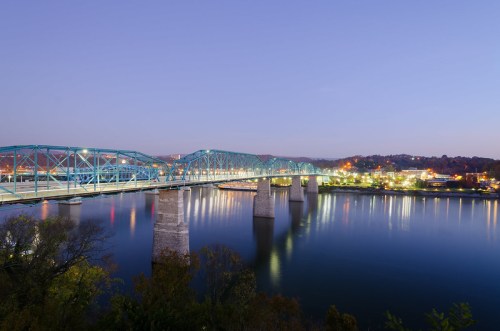 A blue bridge spans a calm river, illuminated by city lights at dusk, with hills in the background.