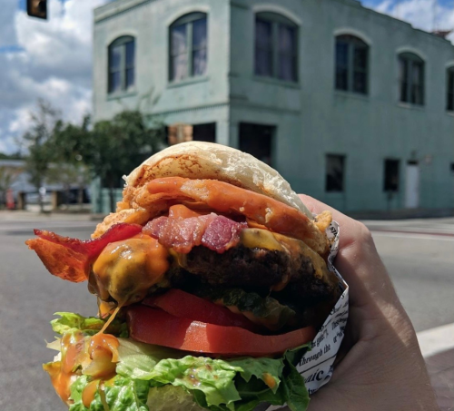 A hand holding a loaded burger with bacon, cheese, lettuce, and tomato, with a vintage building in the background.
