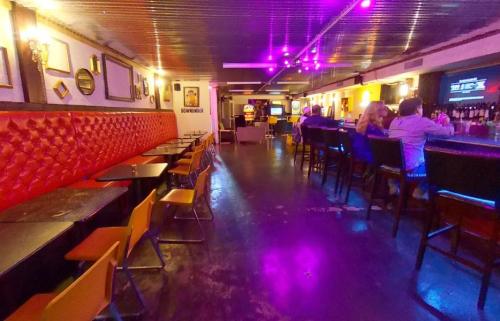 A dimly lit bar with red leather seating, tables, and a long bar area, featuring colorful lights and a lively atmosphere.