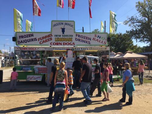A busy food stand at a fair, offering frozen daiquiris, funnel cakes, lemonade, and corn dogs, with people in line.