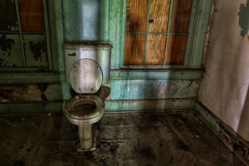 An old, dirty toilet in a dilapidated room with peeling paint and dusty floors. Light filters through boarded windows.