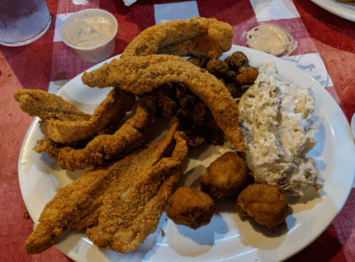 A plate of fried catfish, fried okra, hushpuppies, and a creamy side dish, served on a red-checkered tablecloth.