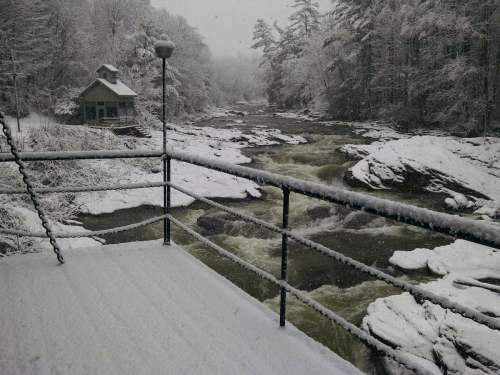 A snowy landscape with a river flowing through, viewed from a deck with a cabin in the background.