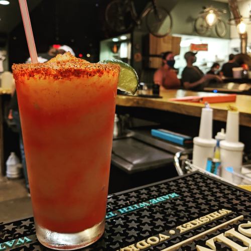 A vibrant cocktail with a chili rim and lime slice, sitting on a bar counter with blurred patrons in the background.