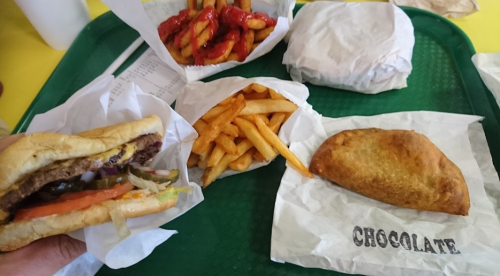 A hand holding a burger next to fries, a chocolate pastry, and a tray with wrapped items.