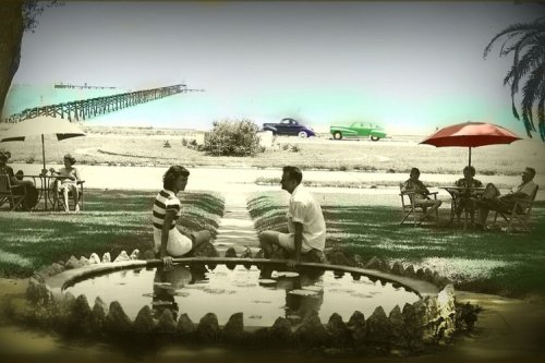 A vintage scene with two people sitting by a fountain, surrounded by palm trees and others relaxing under umbrellas.