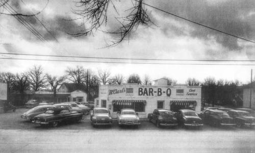 A vintage black-and-white photo of a barbecue restaurant with classic cars parked outside under a cloudy sky.