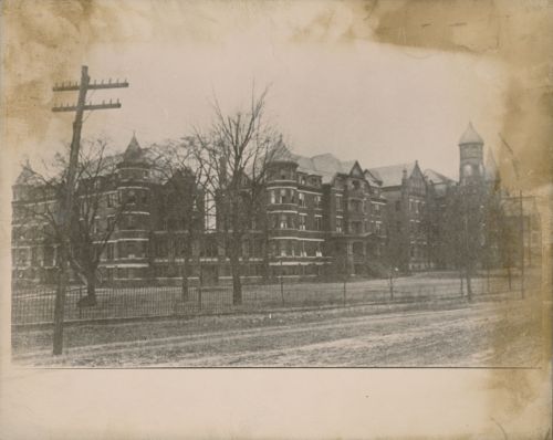 Historic black-and-white photo of a large, old building with a tower, surrounded by trees and a fence.