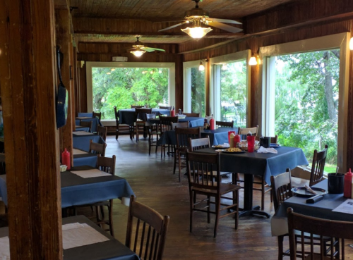 Cozy restaurant interior with wooden decor, tables set with blue tablecloths, and large windows overlooking greenery.