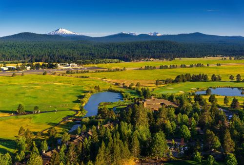 Aerial view of a lush green golf course with ponds, surrounded by mountains and forests under a clear blue sky.