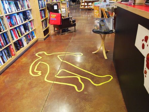 A bookstore interior featuring a chalk outline of a body on the floor, surrounded by bookshelves and a reading chair.