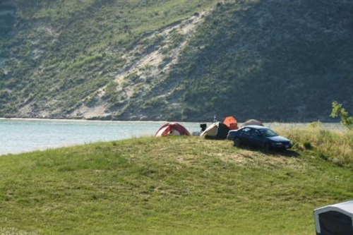 A car parked near tents on a grassy hill by a calm lake, surrounded by green hills in the background.