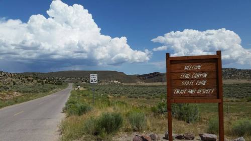 A road leading to a sign for Elmo Canyon State Park, with a speed limit sign and distant clouds in the background.