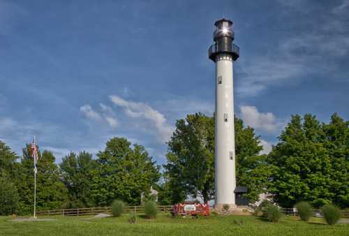 A tall white lighthouse stands against a blue sky, surrounded by green trees and a grassy area.