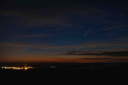 A night sky filled with stars above a small, illuminated town, with a comet visible in the distance.