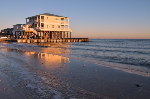 A beach house on stilts at sunset, reflecting on calm waters with gentle waves lapping at the shore.