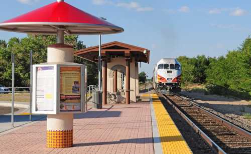 A train approaches a station with a red roof, surrounded by trees and a clear blue sky.