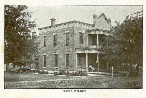 Historic image of Hotel Wilber, a two-story brick building with a porch, surrounded by trees and a grassy area.