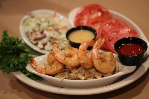 A plate featuring grilled shrimp, shredded chicken, sliced tomatoes, coleslaw, and dipping sauces.