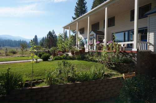 A charming house with a porch, surrounded by lush gardens and mountains in the background under a clear blue sky.