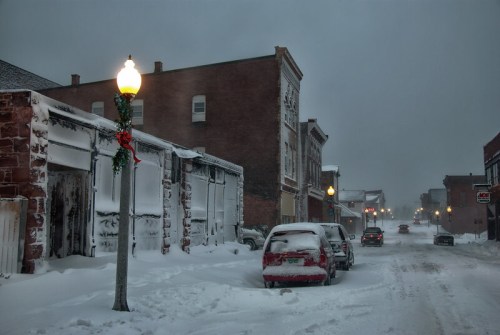 Snow-covered street with parked cars, buildings, and a decorated lamppost in a wintery, overcast atmosphere.