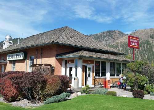 A brick restaurant with a sloped roof, surrounded by greenery and mountains, featuring a sign for "Rivers City Grill."