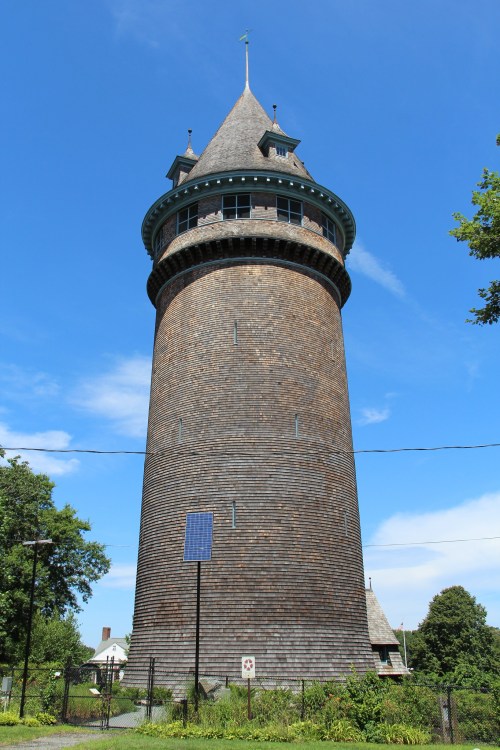 A tall, wooden water tower with a conical roof, surrounded by greenery and under a clear blue sky.
