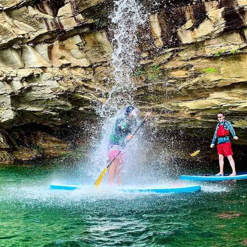 Two people paddleboarding under a rocky overhang, with water splashing down from above.