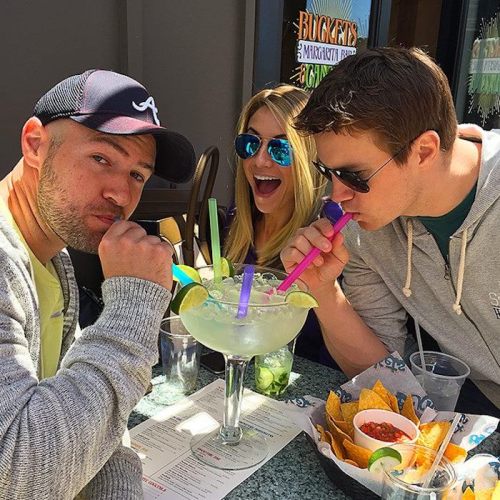 Three friends enjoy a large margarita with colorful straws, sitting at a table with nachos and menus.