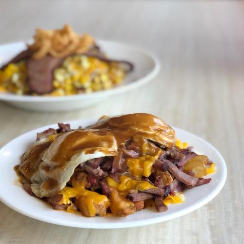 A plate of cheesy hash browns topped with ham, eggs, and gravy, with another dish of corn and meat in the background.