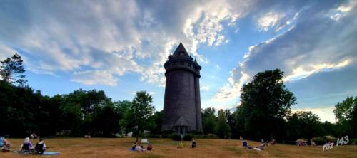 A tall, historic tower surrounded by green trees and people relaxing on the grass under a cloudy sky.